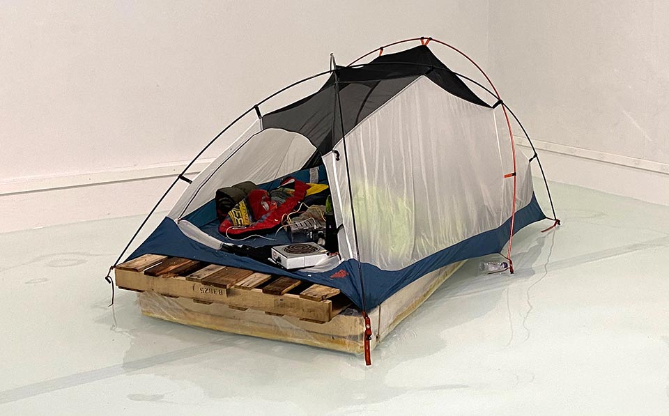 'A floating improvised raft made of plastic sheeting and wooden palettes topped with a small tent'