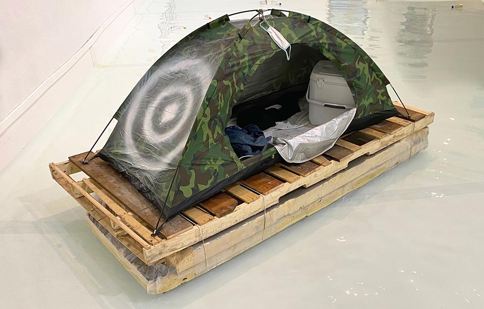 'A floating improvised raft made of plastic sheeting and wooden palettes topped with a small tent. The tent is small and has a target spray painted on one side'